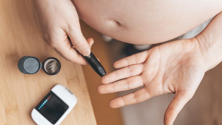 Pessoa grávida monitorando os níveis de glicose no sangue com dispositivo lancetador, destacando a importância do controle do diabetes mellitus e do acompanhamento do diabetes na gestação. Sobre a mesa, podem ser vistos um medidor de glicose e tiras de teste, instrumentos essenciais para o controle do diabetes.
