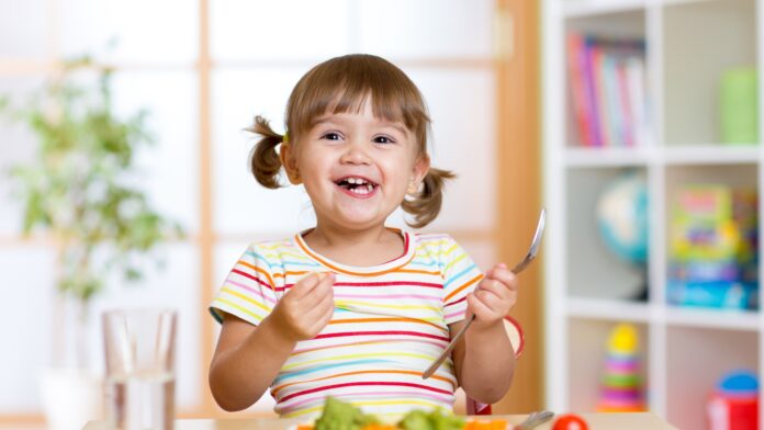 Criança sorridente sentada à mesa segurando um garfo, comendo uma refeição saudável com vegetais ao fundo de um ambiente iluminado e colorido, com estante contendo brinquedos e livros.