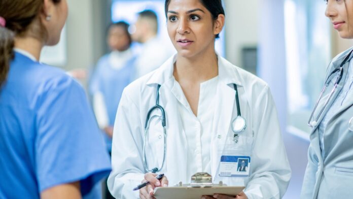 o que é r3? Profissional de saúde usando jaleco branco e estetoscópio, segurando uma prancheta, interagindo com outros membros da equipe em um ambiente hospitalar.