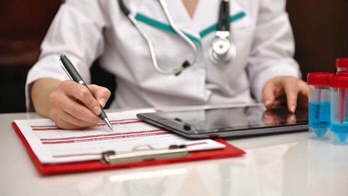 Especialidade médica com pré-requisito Profissional de saúde escrevendo em um formulário médico, com um estetoscópio pendurado no pescoço e materiais de laboratório ao lado.