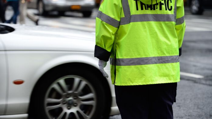 Agente de trânsito com uniforme amarelo fluorescente observando um veículo em movimento em uma via urbana.
