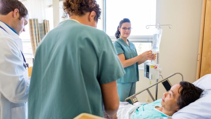 Equipe médica ao redor de uma paciente em um leito hospitalar, com uma profissional ajustando uma bolsa de soro.