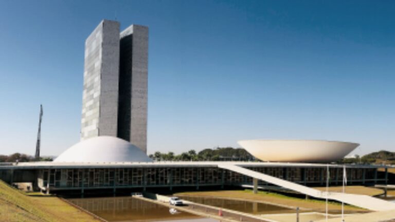 Vista do Congresso Nacional do Brasil, em Brasília, destacando a cúpula do Senado e a base com janelas amplas, refletidas no espelho d'água em um dia ensolarado.