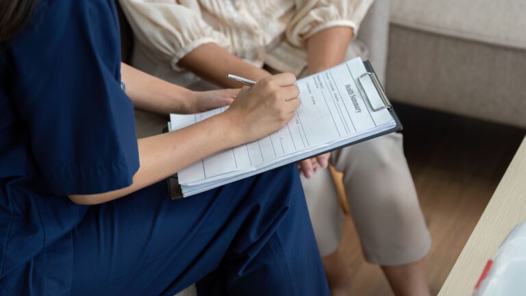 imagem mostra uma profissional da saúde preenchendo um formulário enquanto uma paciete ao lado acompanha.