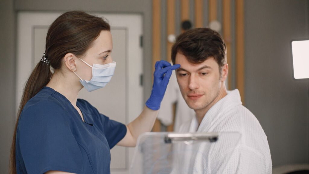 dermatologia clínica | Imagem: Pessoa de pele branca (profissional de saúde) e homem de pele branca (paciente), em plano médio em um consultório clínico ou de estética. À esquerda, a profissional de perfil, de cabelos escuros presos em um rabo de cavalo, veste scrub azul-escuro, máscara cirúrgica descartável clara e uma luva de procedimento azul na mão direita, com a qual toca suavemente a região da testa/supercílio do paciente. À direita, o paciente de cabelos curtos e escuros, vestindo um roupão de banho waffle branco, olha atentamente na direção da profissional enquanto é avaliado. Em primeiro plano, no quadrante inferior central, vê-se o topo de uma prancheta acrílica transparente ligeiramente desfocada. O fundo apresenta uma porta branca e detalhes de decoração em madeira clara e cinza de forma suavemente desfocada.
