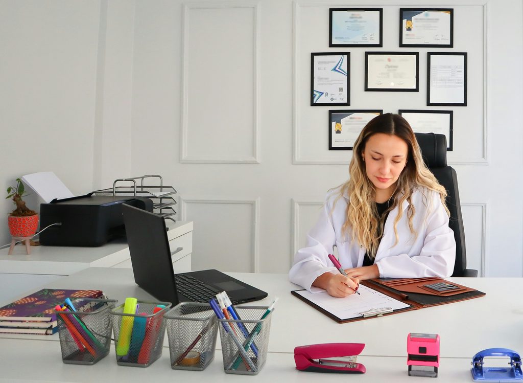 Uma médica branca e loira de jaleco está sentada numa cadeira preta. A sala é branca e tem vários certificados na parede. Ela escreve em uma prancheta e na mesa há um computador, impressora e diversos materiais de papelaria.