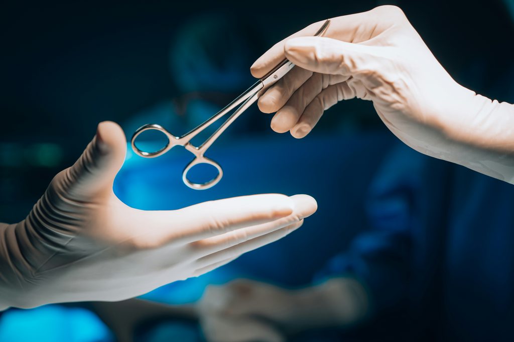 Apenas as mãos de dois médicos aparecem. O fundo é azul por conta da vestimenta comum em centros cirúrgicos. O médico da direita entrega um instrumento cirúrgico para o outro.